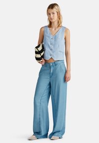 Modrý šálený bezrukávový crop top s výstřihem do V, zdobený knoflíky. V kombinaci s širokými světlomodrými džíny a texturovanými nazouvacími botami.