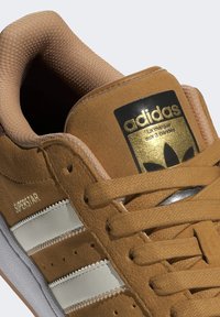 Bruna mockasiner i mocka med tre vita ränder, texturerade snören och en guldig logotag. Rundad tå och kontrasterande sula fulländar designen.