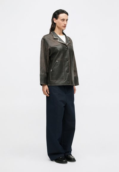 Dunkelbraune Lederjacke mit silbernen Nieten am Kragen und den Taschen; oversized Schnitt und Knopfverschluss, kombiniert mit navyfarbenen Weiten Hosen.