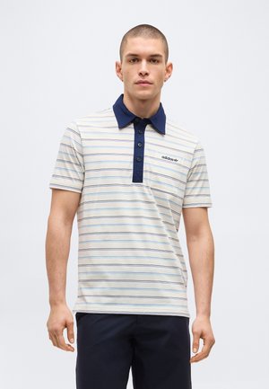 Άνδρας φορώντας λευκή πόλο με κοντά μανίκια και λεπτές μπλε ρίγες, ναυτικό γιακά και πατιλέτα, λογότυπο Adidas στο στήθος και σκούρο παντελόνι.