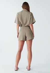 Moteris stovi nugara, vilkėdama alyvuogių žalios spalvos trumpomis rankovėmis marškinėlius ir atitinkančius aukšto juosmens šortus, ant paprasto fono avi white sandalais.