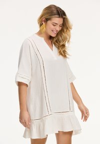 Weißes Kleid mit V-Ausschnitt, kurzen umgeschlagenen Ärmeln, strukturiertem Stoff und horizontalen Ausschnitten. Der Saum verfügt über eine Rüsche.