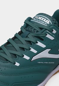 Πράσινο αθλητικό παπούτσι με κορδόνια, λευκό λογότυπο, διάτρητο σύστημα αερισμού και λεπτομέρειες επωνυμίας που περιλαμβάνουν "Joma Maxima" και "Perfect Fit".