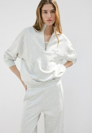 Mujer vestida con sudadera de cremallera parcial de color gris claro con el texto "Tennis Club 1984" y pantalones a juego, de pie con las manos en las caderas sobre un fondo liso.