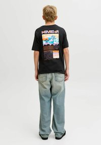 Persoon met kort blond haar, gekleed in een zwart T-shirt met opdruk en losse lichtblauwe spijkerbroek, staand tegen een effen lichte achtergrond.