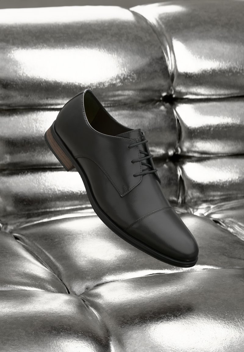 Zapato de vestir de cuero negro con cordones descansando sobre una superficie metálica plateada brillante y acolchada.