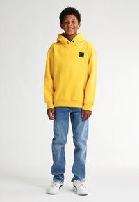 Felpa con cappuccio gialla e logo quadrato, abbinata a jeans azzurri e sneakers bianche. Il tessuto ha una texture morbida.