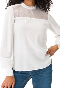 Femme portant un chemisier blanc à manches longues avec un haut de poitrine transparent et un col à volants, assorti à un jean bleu.