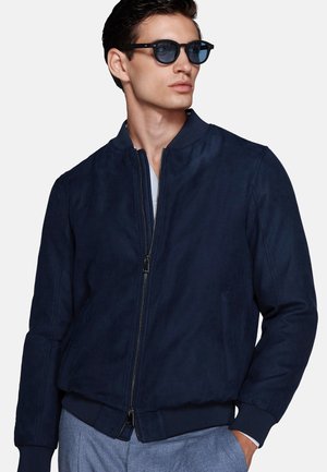 Man wearing dark blue zip-up jacket, light blue pants, and black sunglasses looking to the side with one hand in pocket.