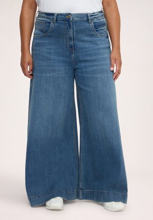 Person iført lyseblå vidde jeans og hvide sneakers, stående mod en ensfarvet baggrund.