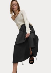 Maglione lavorato a maglia color crema chiaro con spalle scoperte, abbinato a una lunga gonna nera fluente con vita stretta, completato da sandali neri con cinturini.