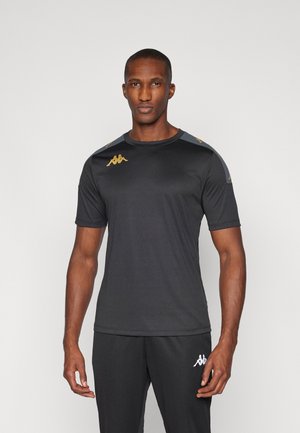 Homme portant un t-shirt de sport noir à manches courtes et un pantalon avec des logos Kappa dorés et blancs sur les épaules, la poitrine et la cuisse, debout devant un fond uni.