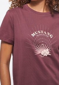Detailní záběr osoby v fialovém tričku s nápisem "MUSTANG true style never dies est. 1932" a růžovým květinovým a tečkovaným kruhovým vzorem.
