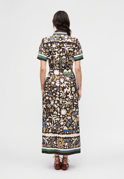Γυναίκα που στέκεται με την πλάτη, φοράει καφέ μακρύ φόρεμα με κοντά μανίκια και πολύχρωμο αφηρημένο και θεματικό τύπωμα με φύση, καθώς και καφέ παπούτσια με κορδόνια.