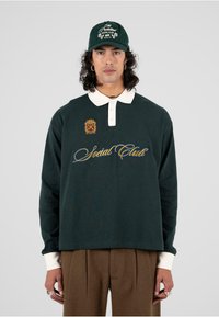 Camisa polo de manga larga verde oscuro con cuello blanco y bordado "Social Club" en dorado y azul. Combinada con pantalones marrones de pinzas y una gorra.