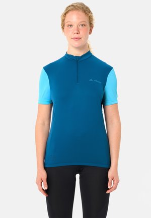 Donna con pelle chiara e capelli biondi ricci indossa una maglia da ciclismo a maniche corte blu e pantaloni da ciclismo neri, in piedi davanti a uno sfondo semplice.