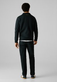 Zwarte sweatshirt met een kraag, gele merklogo op de achterkant, gecombineerd met donkere spijkerbroek en casual schoenen met een blauwe en rode streep.