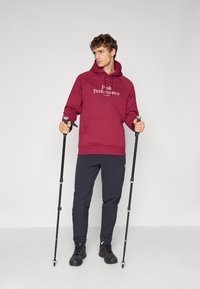 Maroon hættetrøje med logo, snørrebånd og kængurulomme, parret med mørke bukser og sorte vandrestøvler. Brugeren holder trekkingstænger.