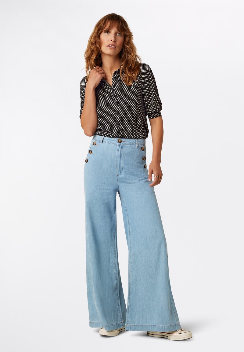 Vrouw staand, gekleed in een zwarte blouse met witte stippen, driekwart mouwen, en lichtblauwe high-waisted wijde jeans met knopen aan de zijkant.