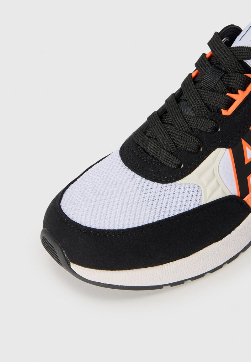 Zapatilla en blanco y negro con parte superior de malla, cordones negros y acentos naranjas en el lateral, mostrada desde un ángulo frontal.