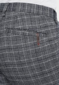 Gebogen grijze plaid broek met een gestructureerde stof, voorzien van een achterzak met een bruine leren accent en een klein rood detail.