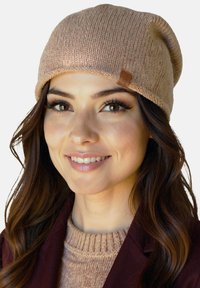 Hosszú barna hajú fiatal nő bézs kötött sapkát, hozzá illő pulóvert és bordó dzsekit visel, miközben mosolyog egy világos háttér előtt.