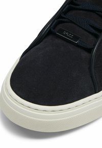 Marinblå mockasneakers med en strukturerad gummisula, vit sömnad och en svart metalllogotyp som accent på snörpanelen.