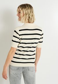 Mujer con cabello corto rubio sonriendo, vistiendo un suéter crema de manga corta con rayas horizontales negras y jeans gris claro, mirando de lado.