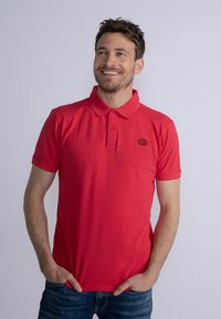 Červené polo tričko s límečkem, krátk�ými rukávy a malým černým logem na hrudi. Nosené s modrými džíny. Hladká textura, volný střih.