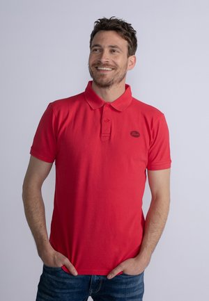 Polo shirt rouge avec col, manches courtes et un petit logo noir sur la poitrine. Porté avec un jean bleu. Texture lisse, coupe décontractée.