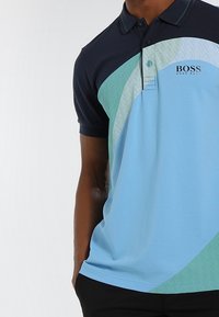 Pikétröja med ljusblå bas och mörkblå ärmar, gröna och teal abstrakta mönster, två knappar och en logotyp på vänster bröst.