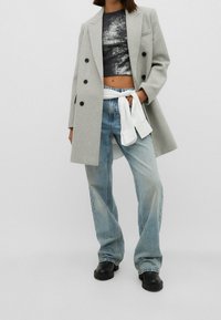 Vrouw met een grijze dubbelgeknoopte jas, zwart metallic cropped top, lichtblauwe jeans met brede pijpen, zwarte laarzen en een wit overhemd dat in de taille is gestrikt.