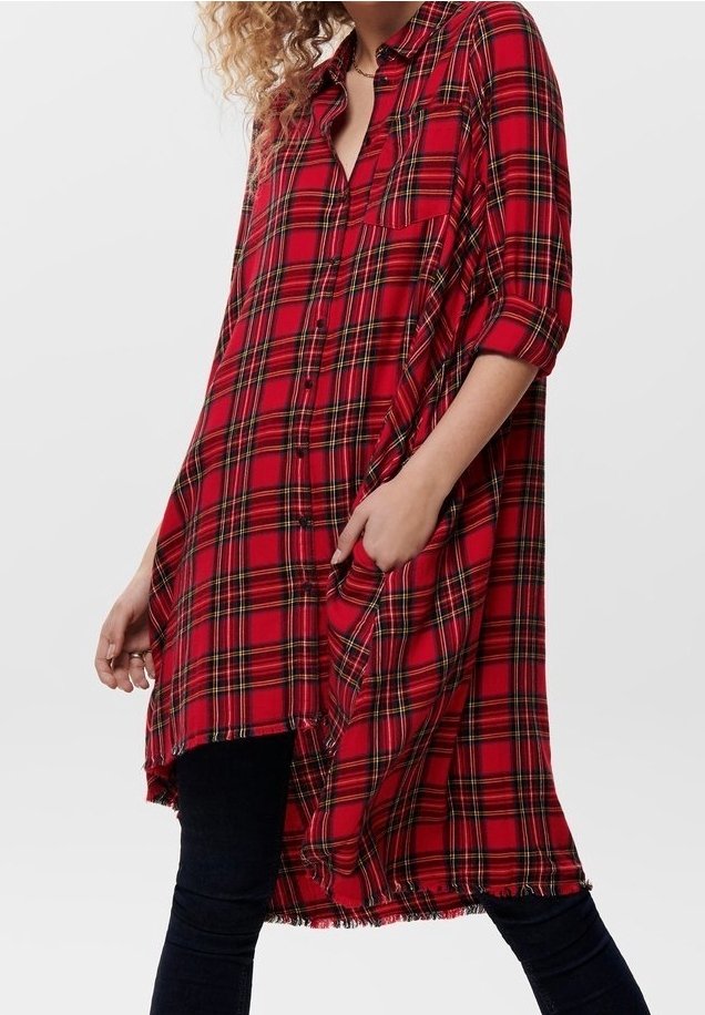 Femme portant une longue chemise-robe à carreaux rouges et noirs avec manches retroussées et pantalon noir, main dans la poche, sur fond blanc uni.