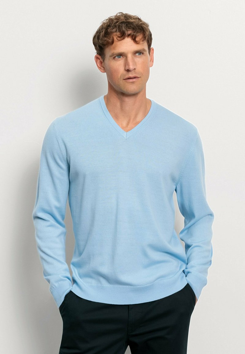 Hellblauer V-Ausschnitt-Pullover aus weichem Stoff, mit langen Ärmeln und gerippten Bündchen. Über einer dunklen Hose getragen, auf einem neutralen Hintergrund drapiert.