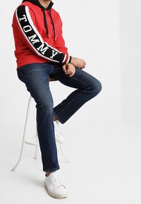 Czerwona bluza z kapturem z czarnymi akcentami i logo "TOMMY" na rękawie, w połączeniu z ciemnoniebieskimi dżinsami i białymi trampkami, siedząca na białym stołku.