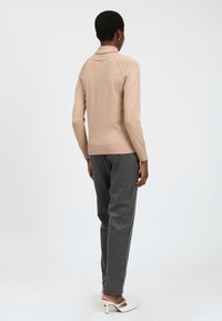 Maglione beige a coste con colletto alto e una lucentezza sottile, abbinato a pantaloni grigi su misura. Dettaglio del logo dorato ben visibile sul retro.