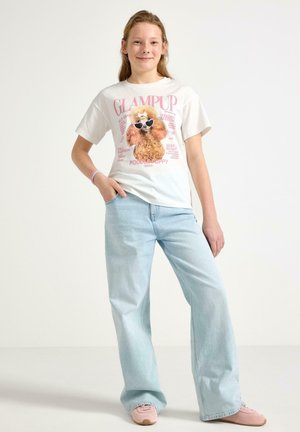 Ung person står med hånden i lommen iført hvid t-shirt med pudelmotiv, lyseblå vidde-bukser og lyserøde sneakers.