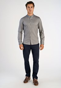 Un giovane uomo sta in piedi di fronte indossando una camicia grigia senza colletto e con bottoni, pantaloni scuri e scarpe stringate marroni su uno sfondo neutro.