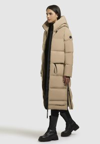 Cappotto lungo beige in stile puffer con cappuccio, dotato di dettagli neri delle zip e lacci, abbinato a stivali militari neri con lacci.