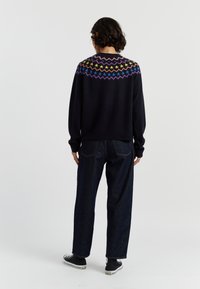 Zwarte sweater met kleurrijke sterren- en zigzagpatronen langs de schouderpartij, gedragen met donkerdenim jeans en zwarte sneakers. Zacht materiaal, ontspannen pasvorm.