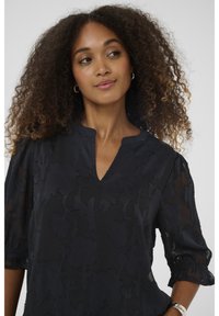 Melnā blūze ar ziedu rakstu, ar V formas kakla izgriezumu, caurspīdīgu materiālu, 3/4 garuma zvanu piedurknēm un smalkiem tekstūras akcentiem visā audumā.