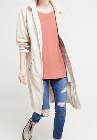 Beige wasserfester Mantel mit Seitentaschen, getragen über einem korallenfarbenen Oberteil und verwaschenen blauen Jeans. Verfügt über einen klassischen Kragen und eine Knopfverschluss.