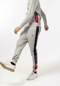 Henkilö, joka käyttää harmaita verkkareita, joissa on tummansinisiä ja punaisia raitoja, sekä U.S. Polo Assn. -logoa ja valkoisia tennareita, astuu eteenpäin.