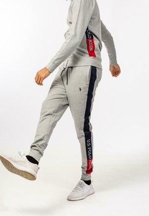Person iført grå sweatpants med marineblå og røde striber, U.S. Polo Assn. logo og hvide sneakers, der træder frem.