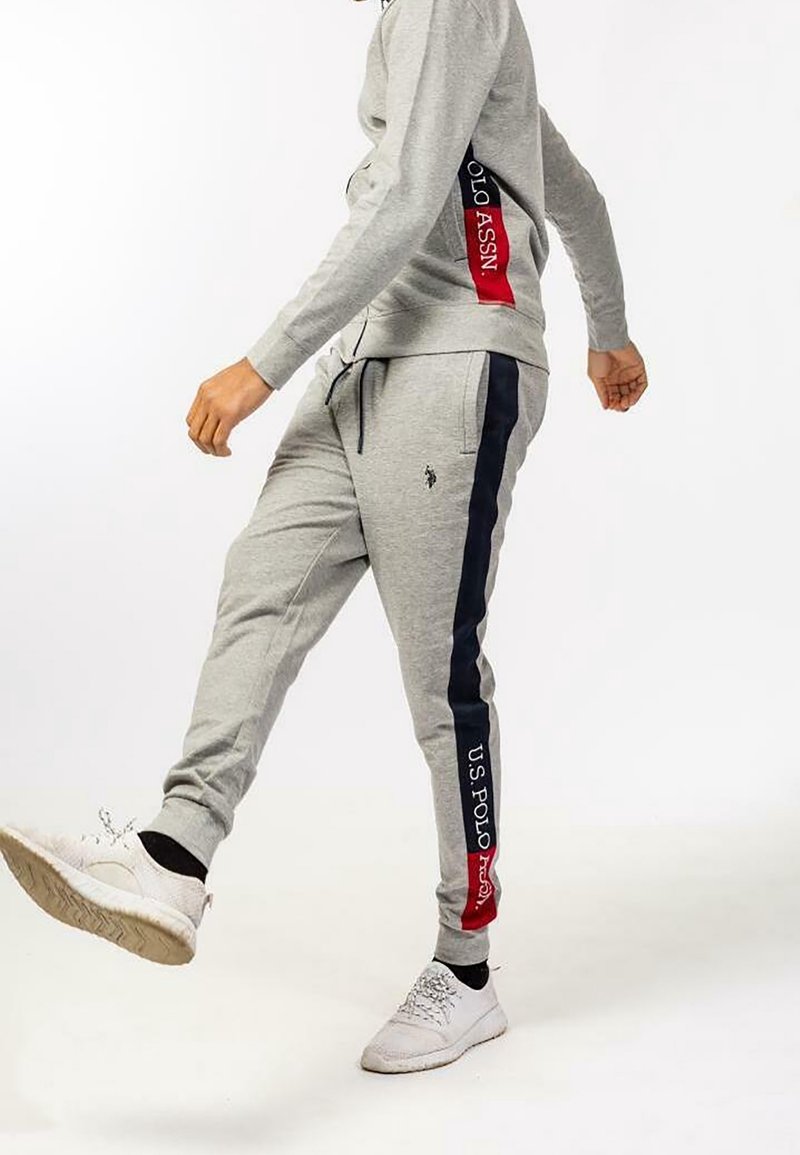 Henkilö, joka käyttää harmaita verkkareita, joissa on tummansinisiä ja punaisia raitoja, sekä U.S. Polo Assn. -logoa ja valkoisia tennareita, astuu eteenpäin.