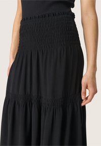 Mujer con un vestido negro de cintura fruncida y secciones de falda, de pie con una mano relajada a su lado contra un fondo neutro.