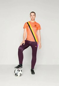 Oranssi urheilupaita, jossa on musta ja keltainen viisto raita ja logo, yhdistettynä purppuraan legginsiin ja mustiin urheilukenkiin, seisoo jalkapallon päällä.