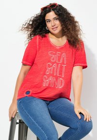 T-shirt rouge à manches courtes avec le texte blanc "SEL SABLE MER", présentant un décolleté arrondi et de petits détails de boucle sur le côté. Porté avec un jean bleu.