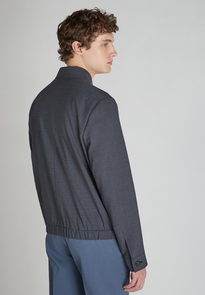 Czarna bomberka o subtelnej fakturze, z elastycznym dołem i zapinanymi na guzik mankietami, noszona na jasnoniebieskich spodniach, widok z tyłu.