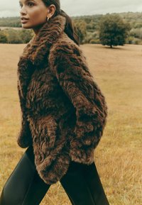 Brūns mākslīgā kažokāda ar plašu apkakli, teksturētu virsmu un sānu kabatām, kombinēta ar melniem pieguļoņiem biksēm dabiskā āra vidē.